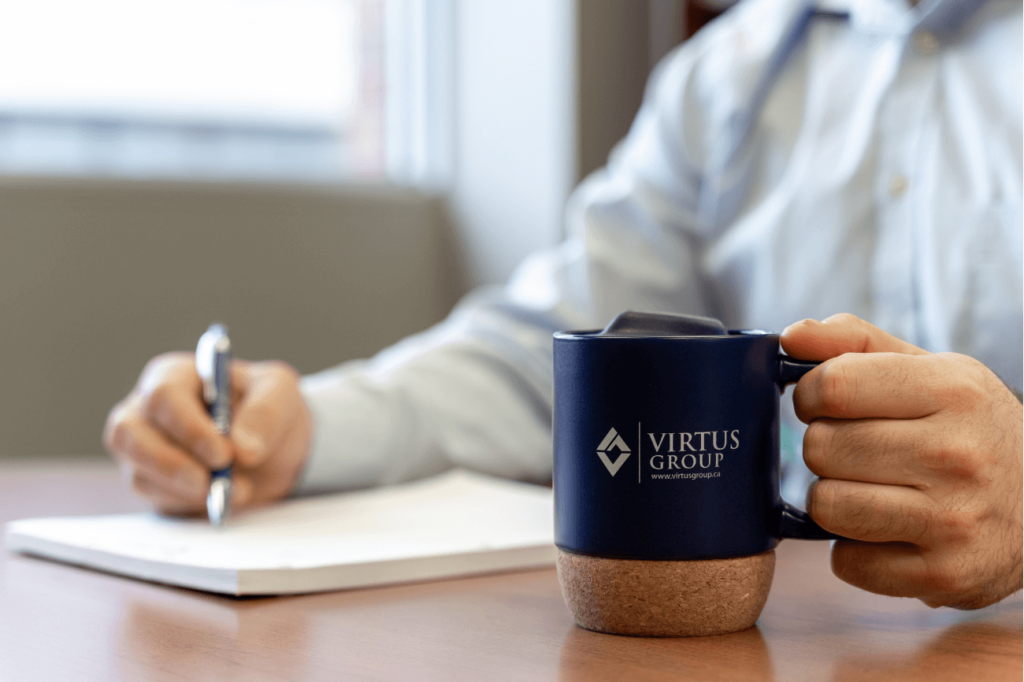 Man in dress shirt holding a blue Virtus Group LLP mug and writing with pen on pad of paper