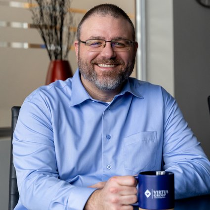 Man with beard in glasses and blue shirt holding blue Virtus Group coffee mug and smiling at camera