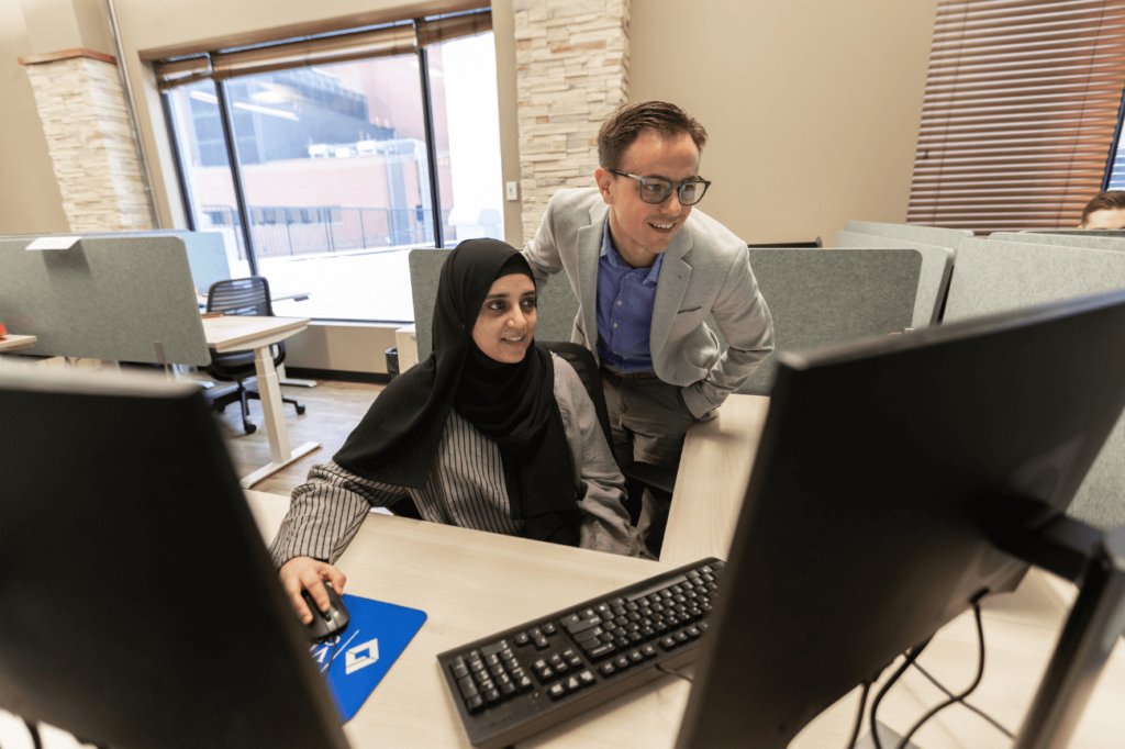 Smiling woman in hijab sitting in front of computer with man in grey blazer and glasses standing behind smiling